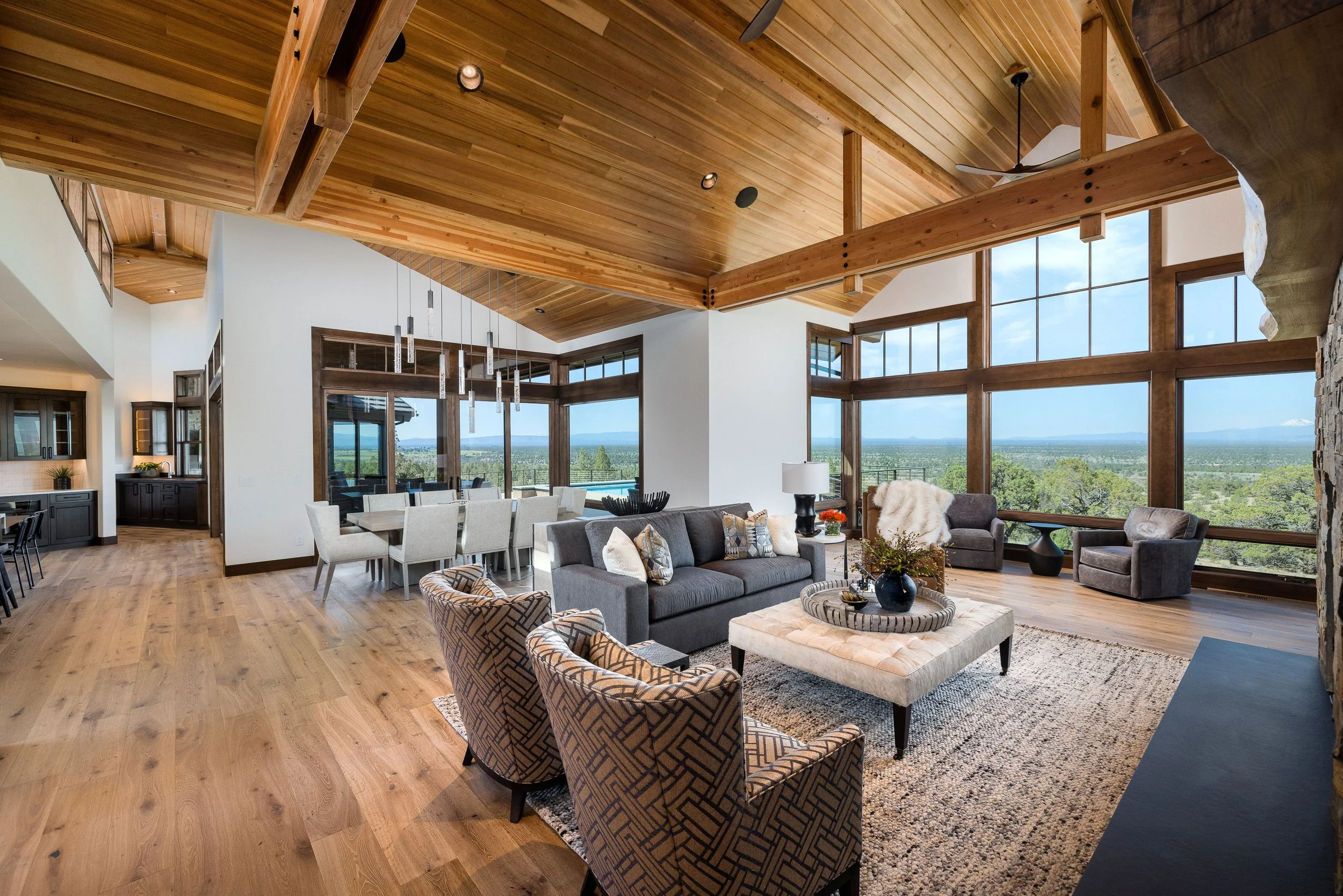 Open living room with wooden ceiling, large windows showing blue sky and green landscape, modern furniture including gray sofas, patterned armchairs, and a coffee table with a black vase and flowers.