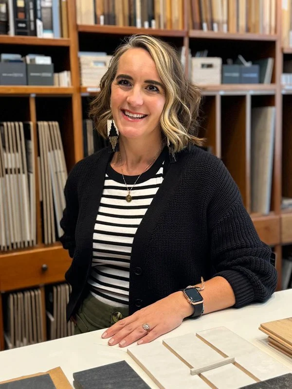 A woman with shoulder-length wavy blonde hair, wearing a black cardigan over a striped black and white top, smiling and sitting at a table in a library or office with wooden shelves filled with books and samples behind her.