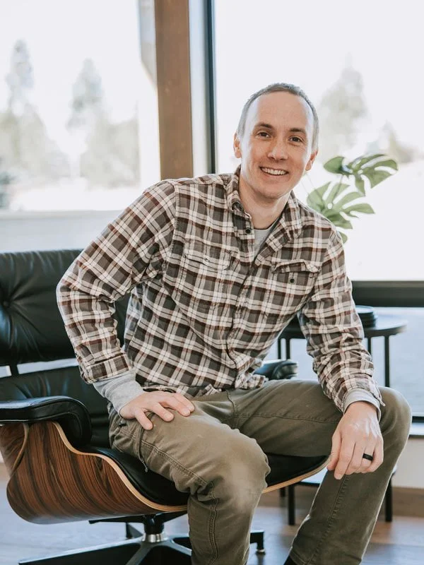 A man sitting on a modern black and wood chair in a bright room with large windows and a green plant.