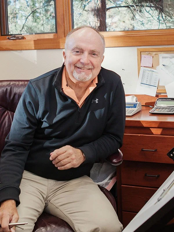 Smiling middle-aged man sitting in an office with a window, desk, and bulletin board behind him.