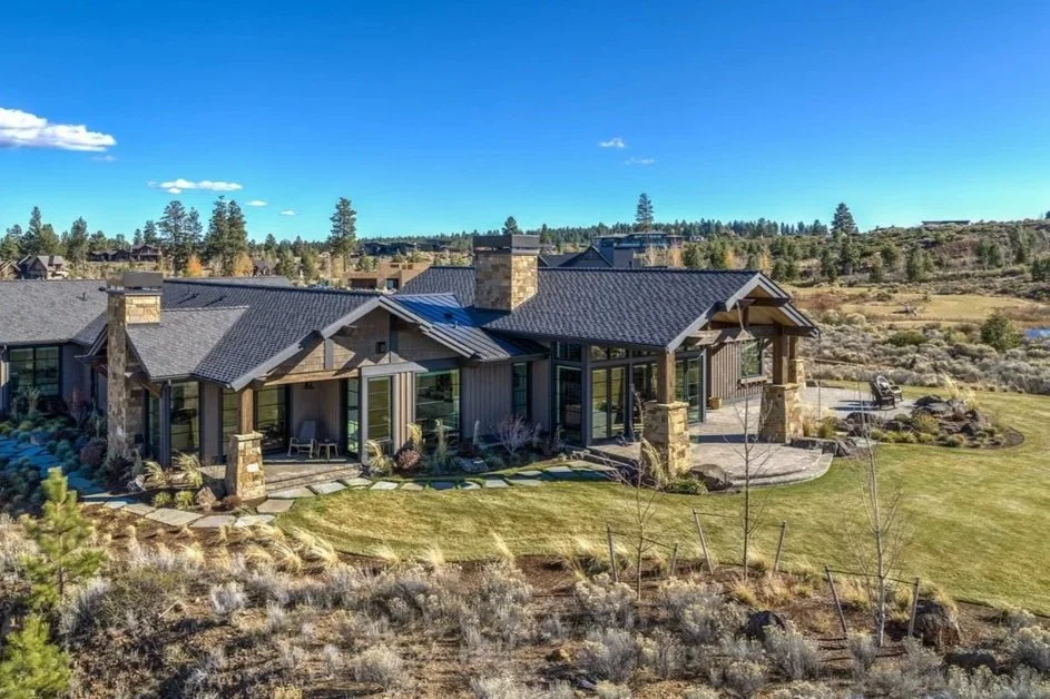 A large modern house with a dark roof, stone pillars, and glass windows, surrounded by a well-maintained lawn and a scenic landscape with trees and hills in the background.