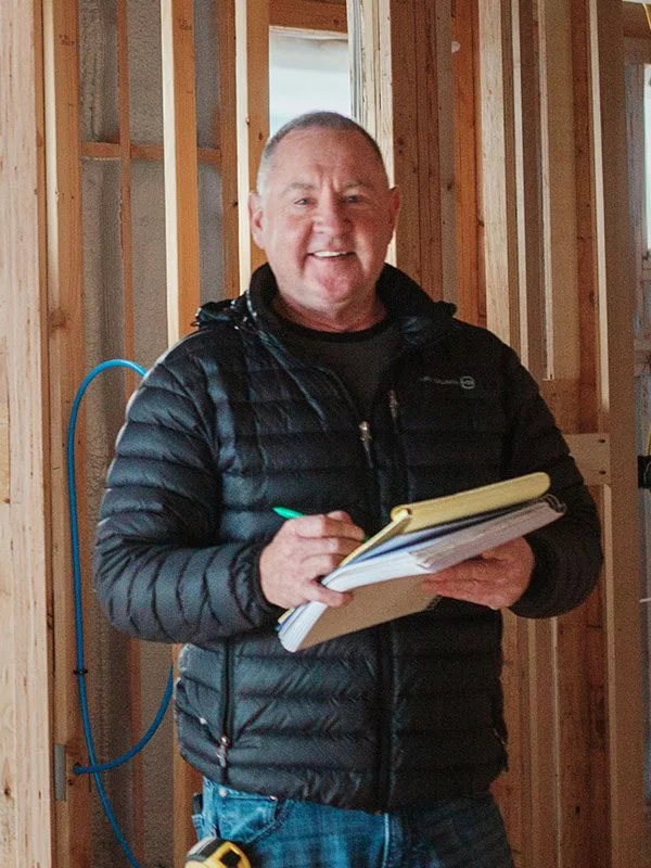 Man in black puffer jacket holding a notebook and pen inside a wooden framing of a building.