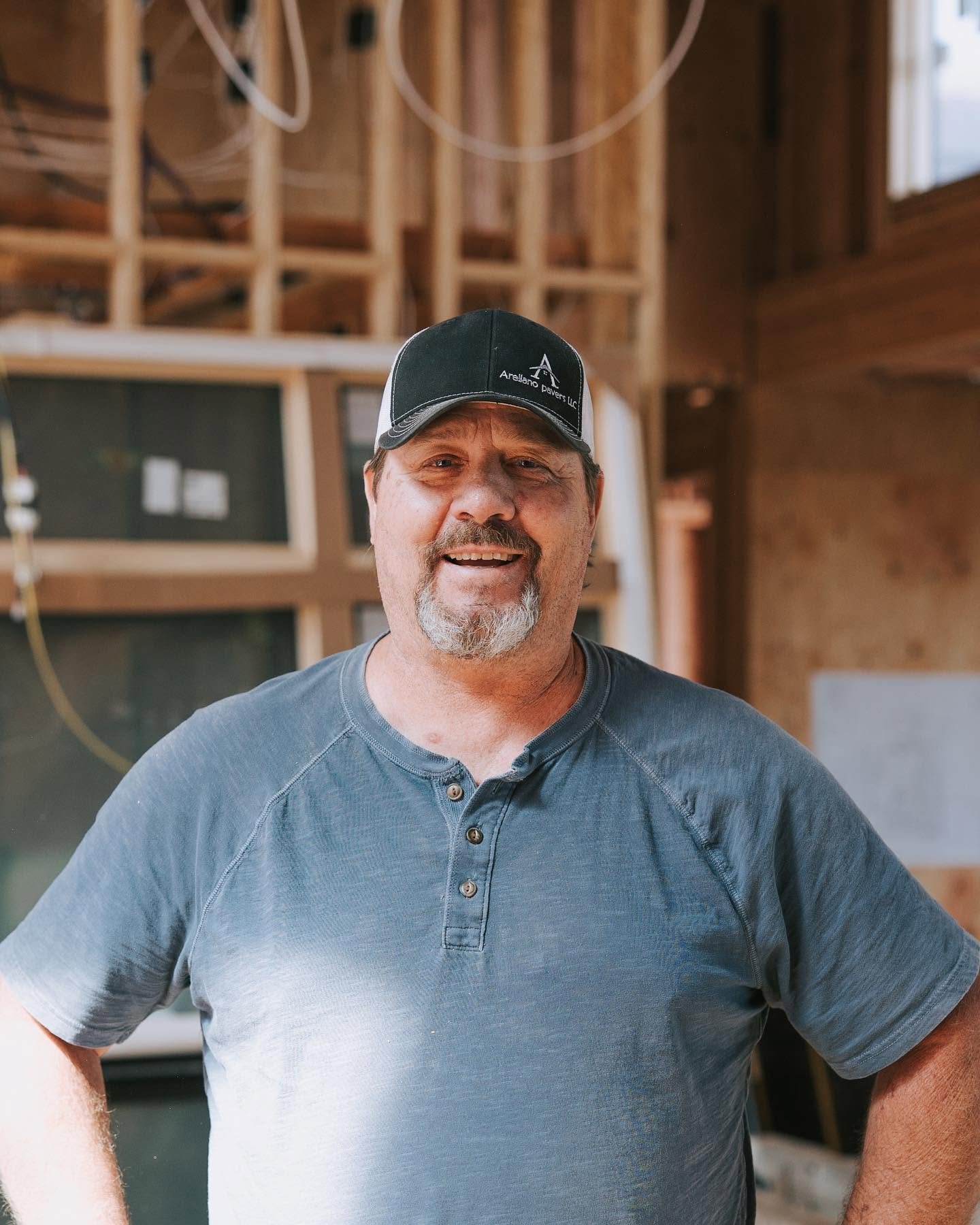 Man wearing a black cap and a gray Henley shirt, smiling inside a wooden construction site.