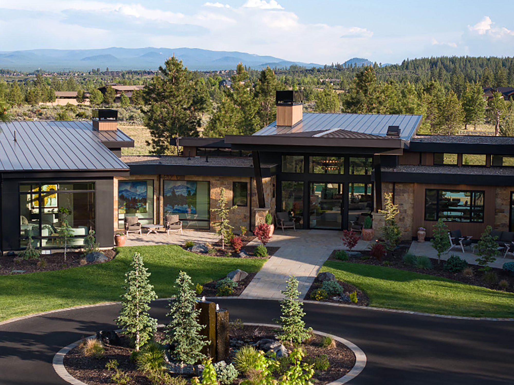 The image shows a modern house with large glass windows, a stone facade, and a metal roof. It has a landscaped front yard with small trees, shrubs, and a pathway leading to the entrance. In front of the house, there is a roundabout with a garden in the center, decorated with young pine trees and flowers. In the background, there are dense forested hills and mountains under a blue sky with some clouds.