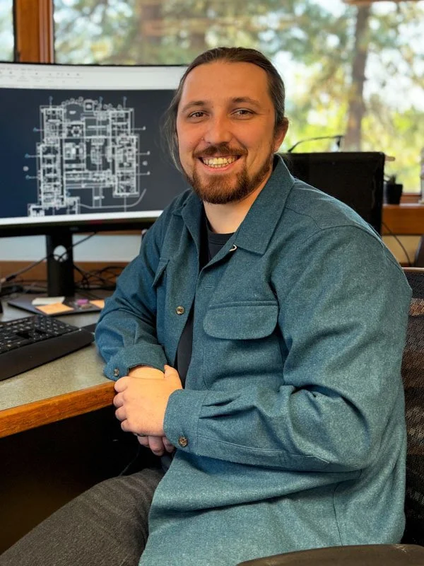 A man with brown hair and a beard smiling at the camera, seated at a desk in front of a computer monitor displaying architectural blueprints, with a window and green scenery in the background.