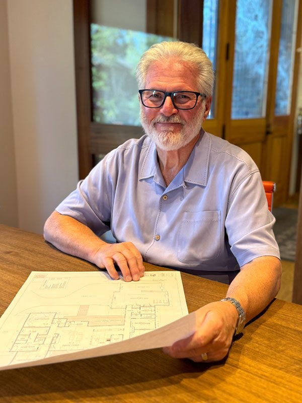 An elderly man with glasses and a beard sitting at a wooden table, holding a set of blueprints or architectural plans.