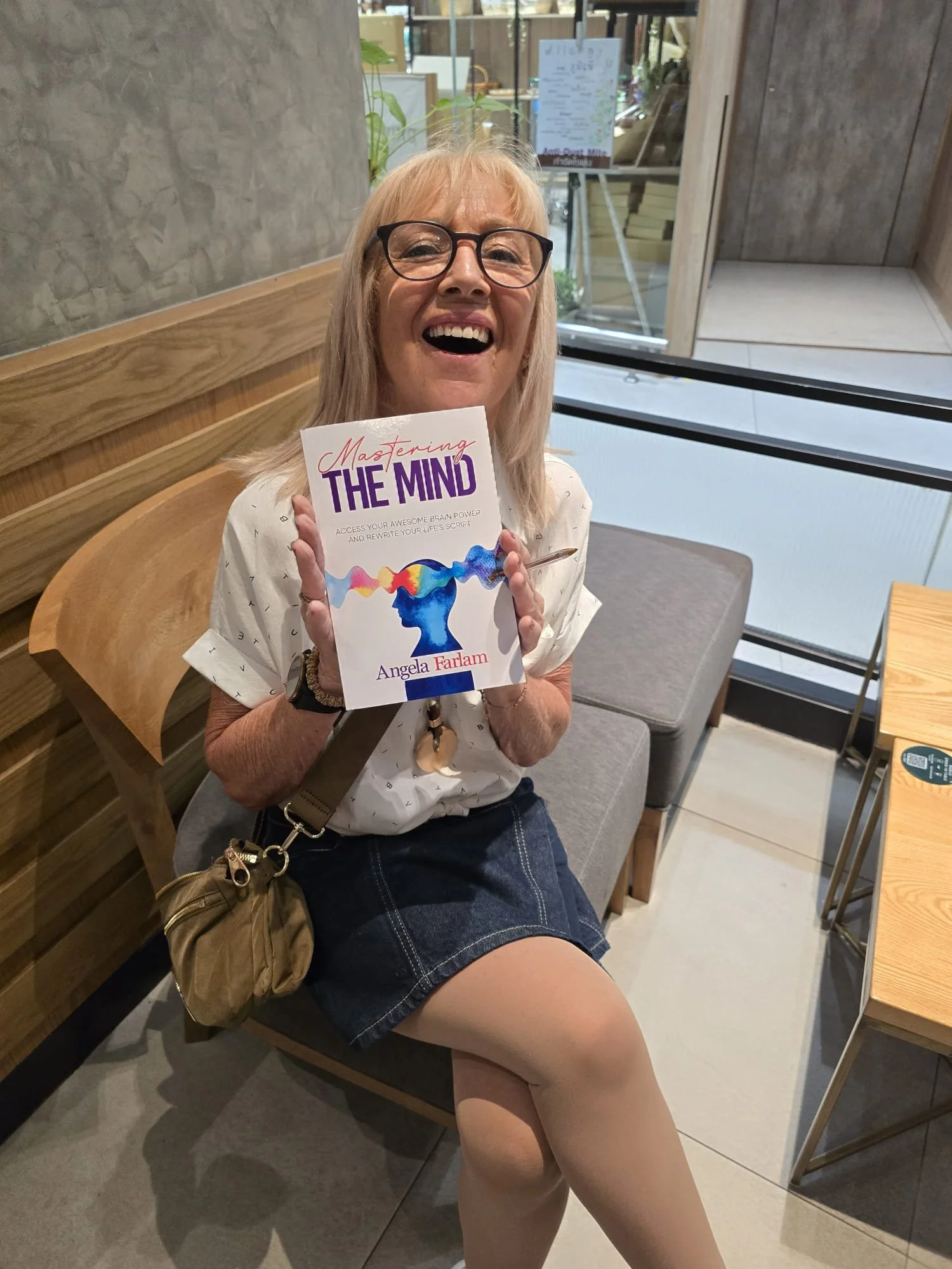 A woman with blonde hair and glasses smiling while holding a book titled "Mastering the Mind" in a casual indoor setting with wooden furniture and large windows.
