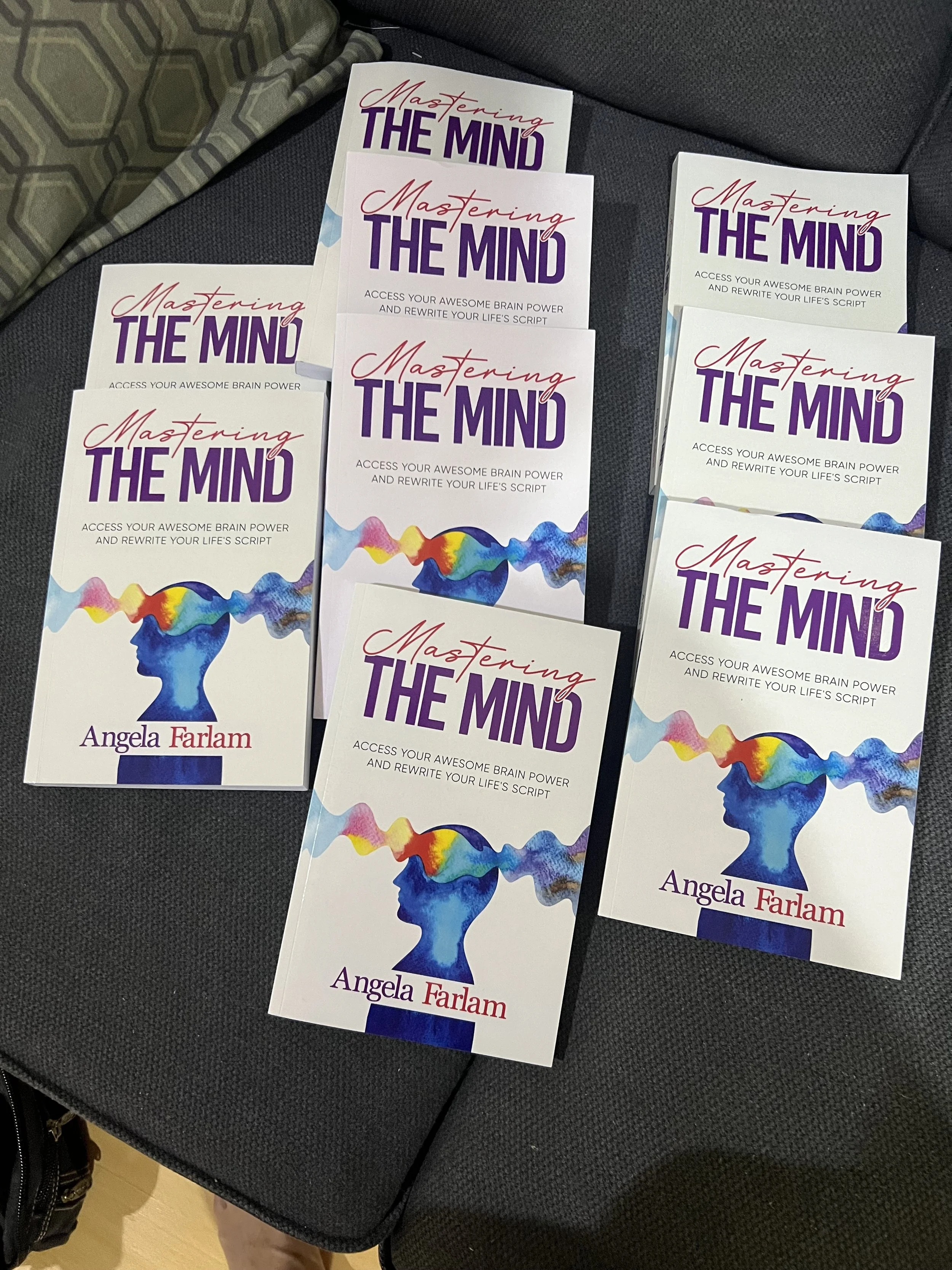 Multiple copies of a book titled "Mastering the Mind" by Angela Farlam are laid out on a black textured surface. The book cover has a white background with purple and red text and a colorful illustration of a human head with a brain made of rainbow-colored splashy patterns.