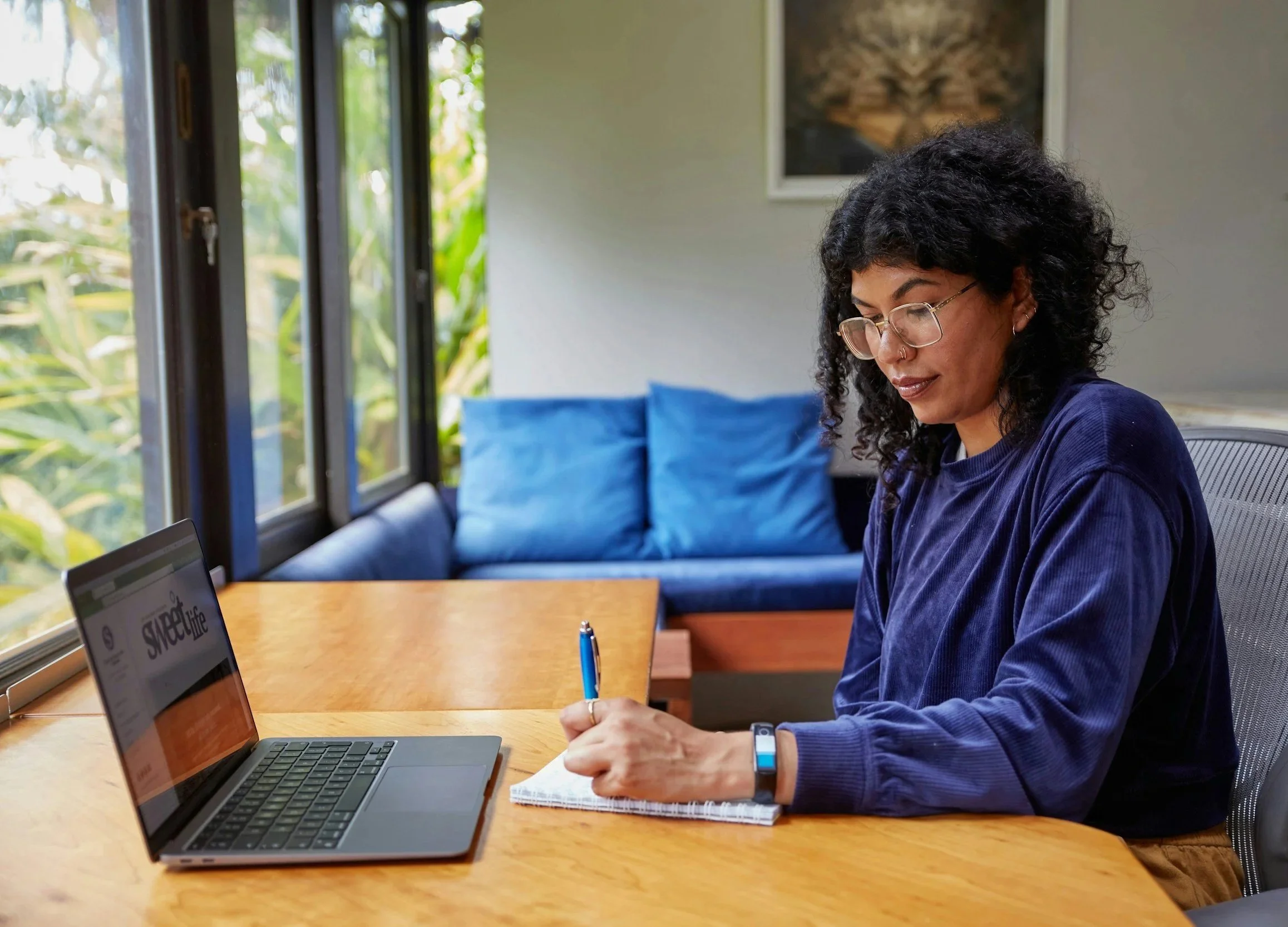 A woman with curly hair, glasses, and a nose ring, sitting at a wooden table with a laptop, a notebook, and a pen, in a room with large windows and a blue couch in the background.