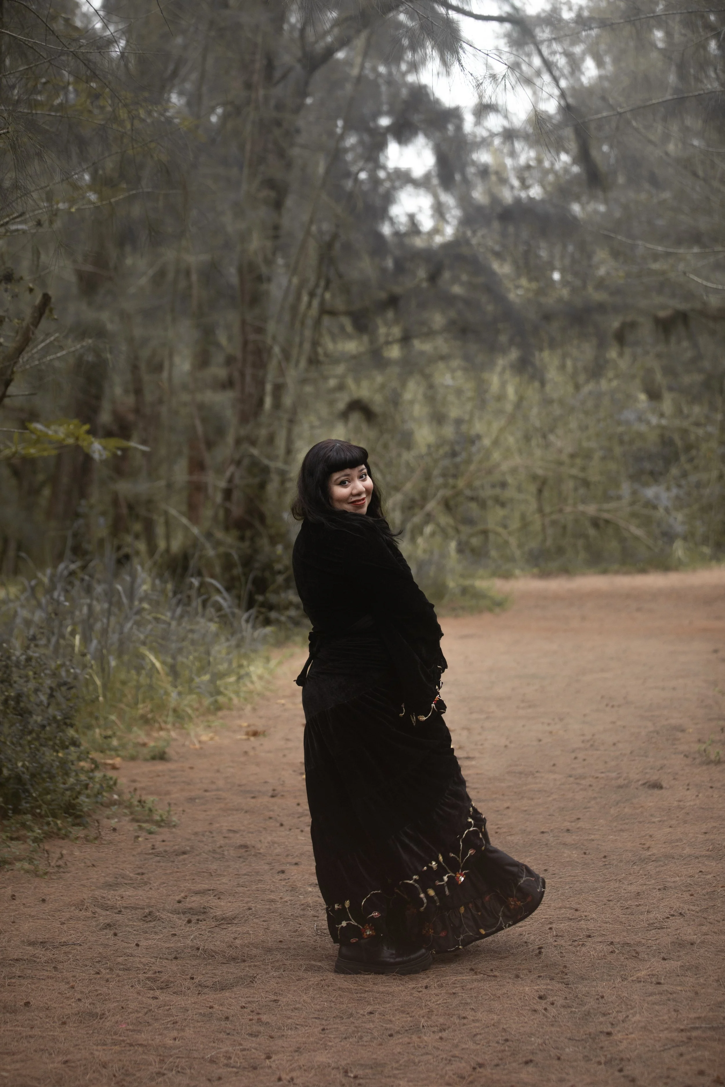 Una mujer con vestido negro y zapatos negros posa en un camino de tierra en un entorno boscoso con árboles altos y ramas en el fondo.