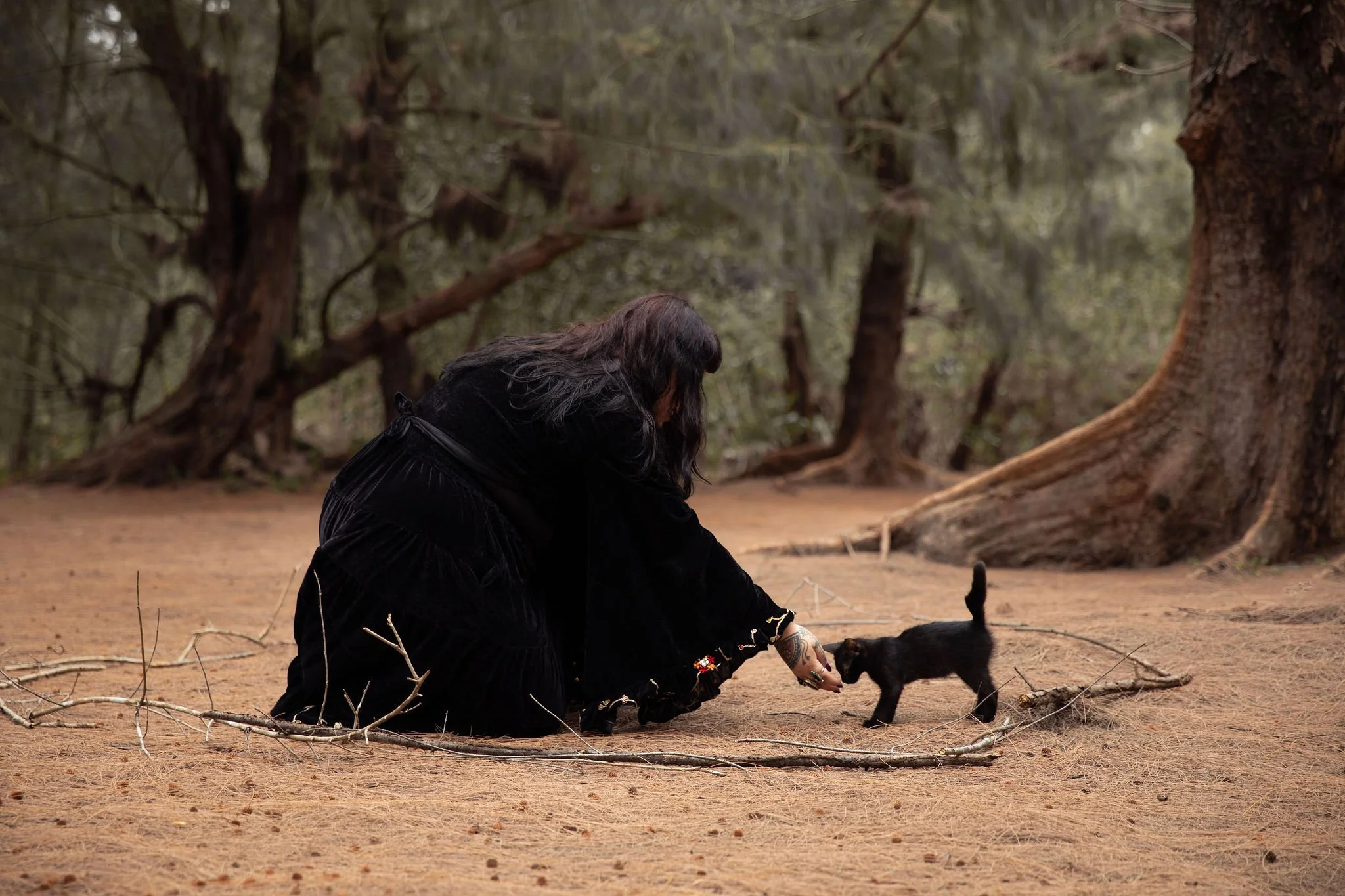 Una mujer con cabello largo y oscuro, vestida con ropas negras, está agachada en un bosque, sosteniendo un pequeño gato negra con cola en alto.