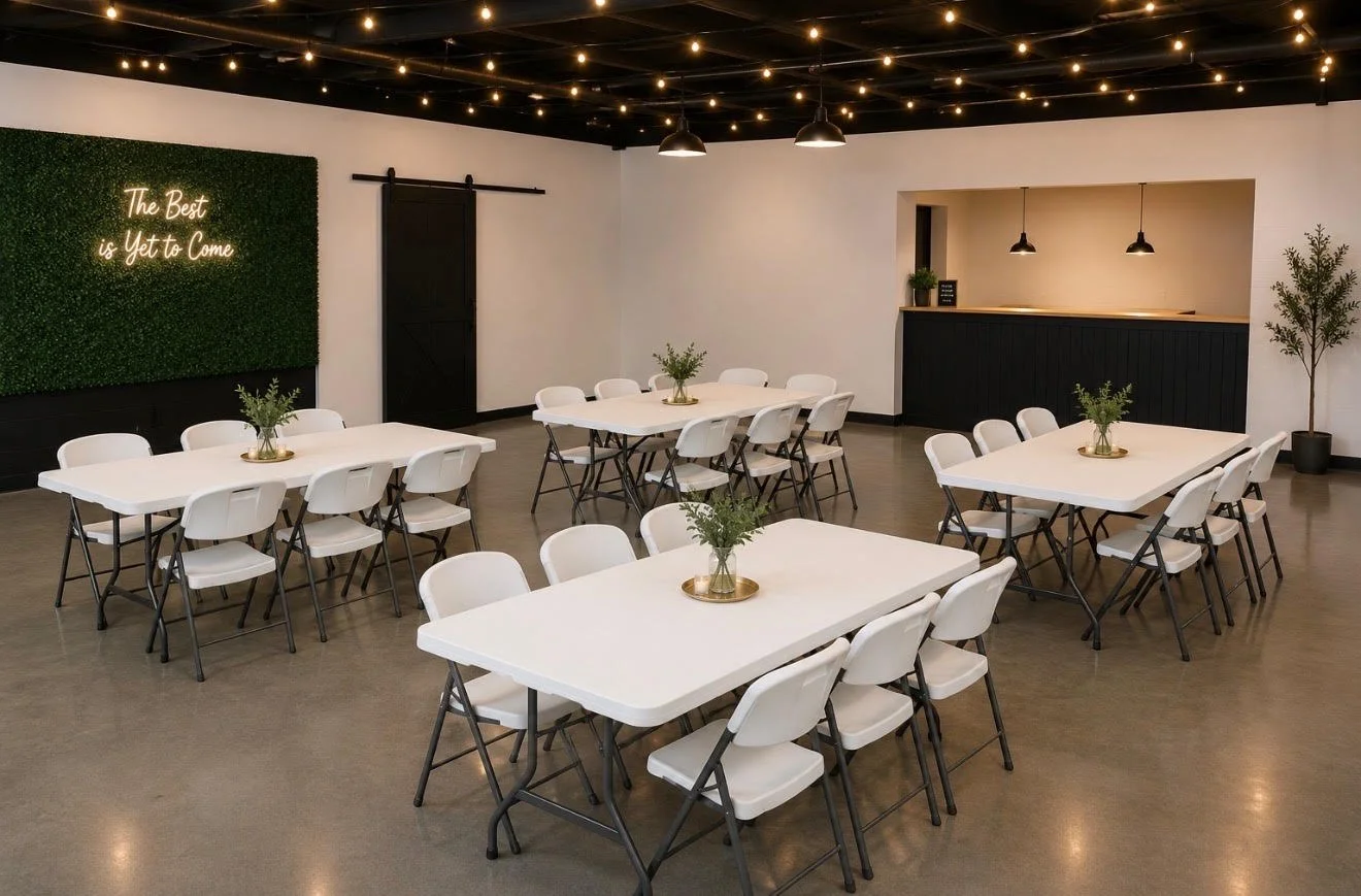 Interior of a modern event space with white tables, white chairs, green plants, black pendant lights, string lights on the ceiling, and a green wall with a neon sign that reads 'The Best is Yet to Come.'