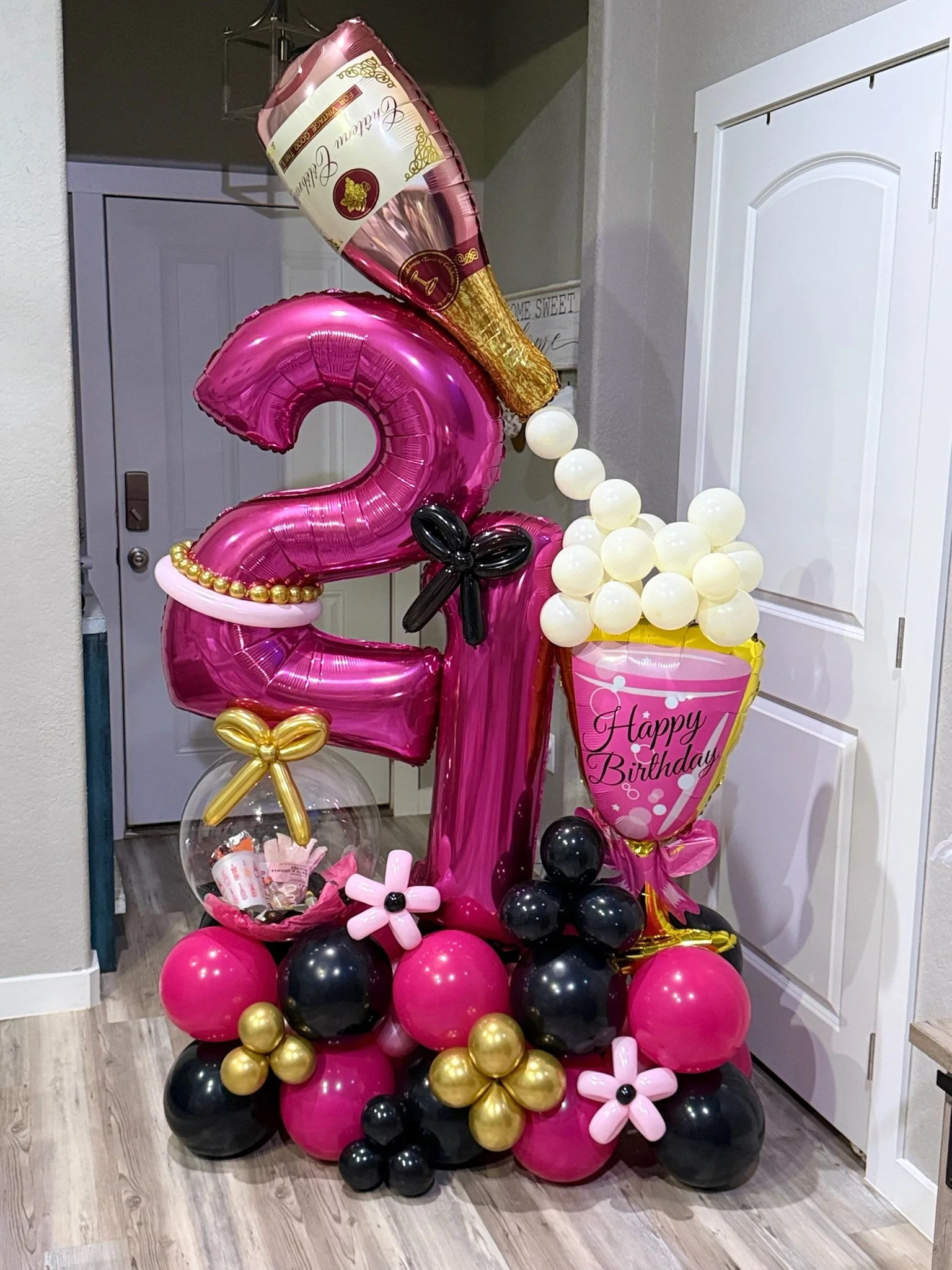 Balloon arrangement for a 20th birthday celebration, featuring a large pink number 20 balloon, gold and black balloons, balloon flowers, and a cone-shaped balloon with a wine bottle design, placed by a door and on a wooden floor.