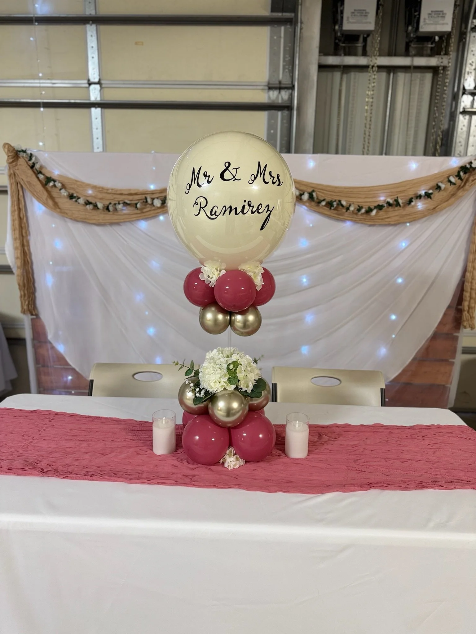 Decorative wedding table centerpiece with a large balloon inscribed "Mr & Mrs Ramirez," surrounded by pink and gold balloons, white flowers, and candles, with a wedding backdrop decorated with draped fabric and lights.