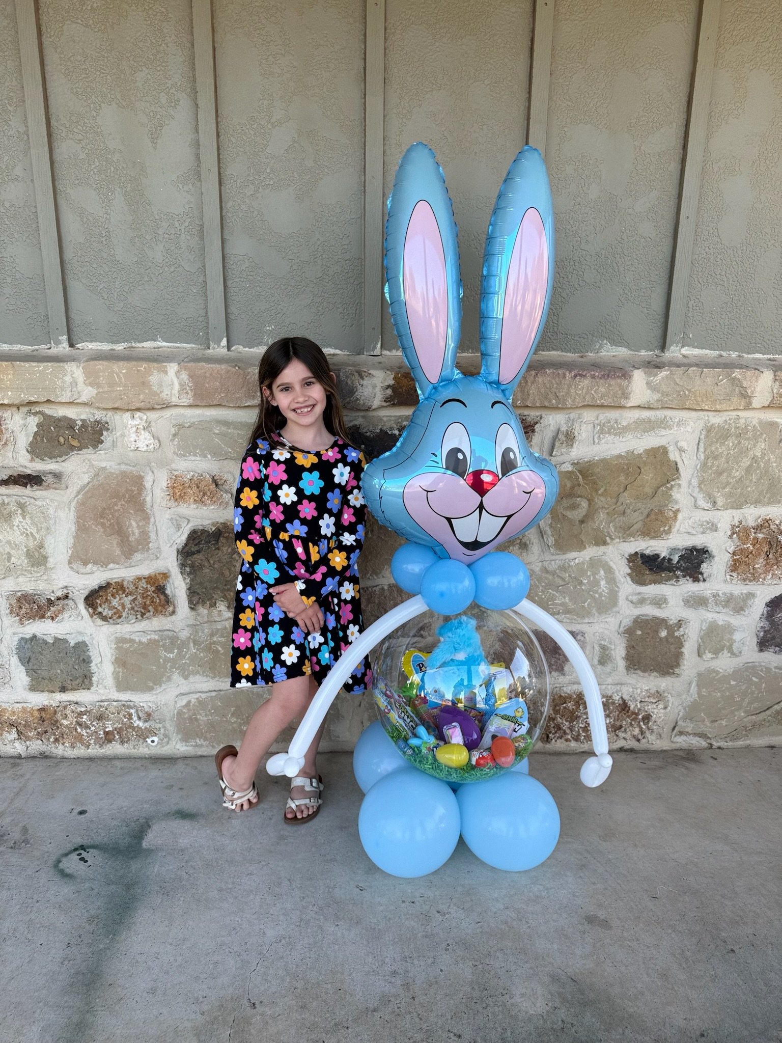 A young girl wearing a black dress with colorful flowers standing next to a large blue Easter bunny shaped balloon decoration. The decoration contains a transparent container with plastic eggs inside. The girl is smiling and standing outdoors against a stone and stucco wall.
