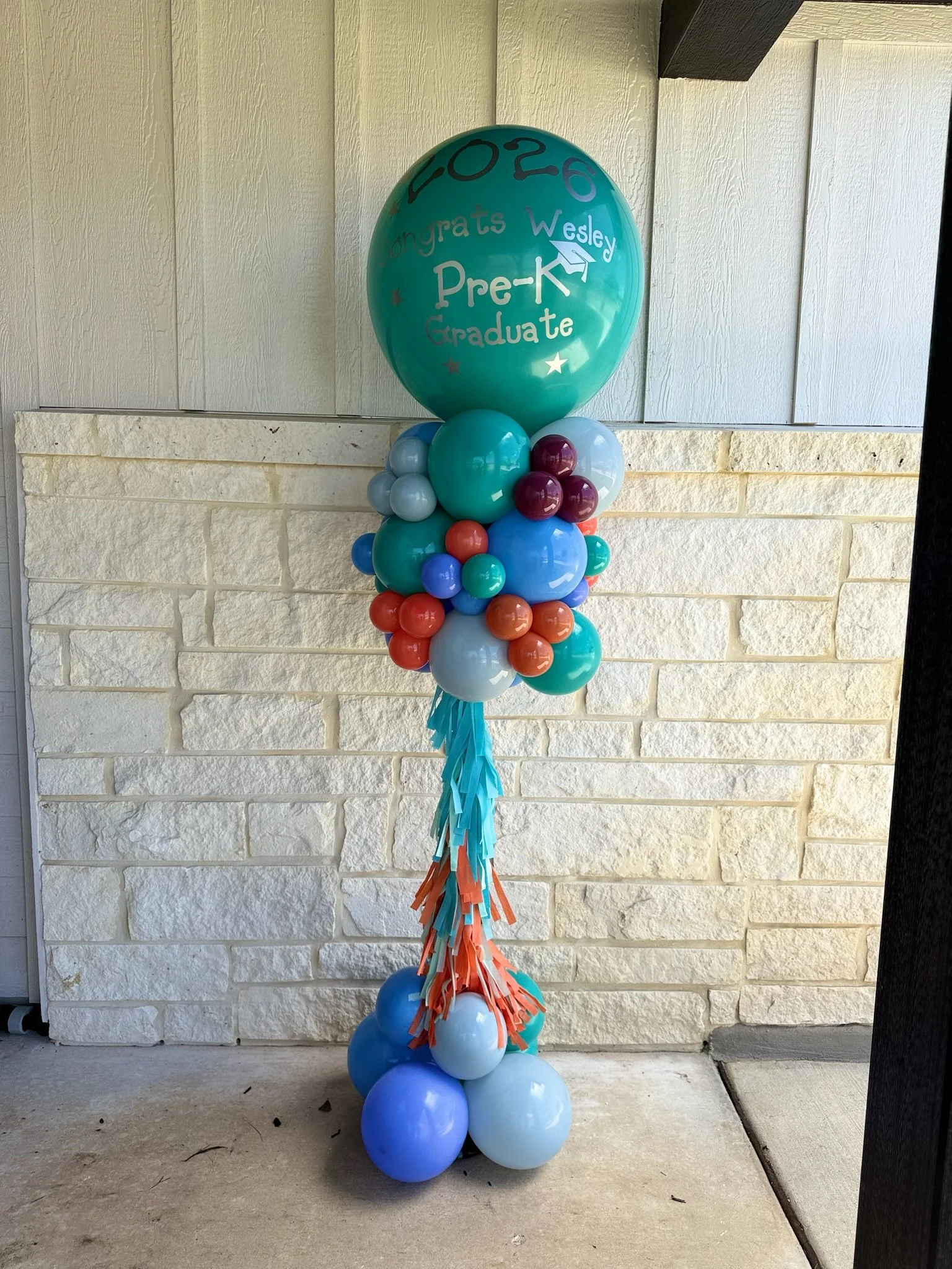 A balloon bouquet with a large teal balloon that says 'congrats Wesley Pre-K Graduate' in white, surrounded by smaller blue, teal, purple, and orange balloons, with hanging streamers in matching colors.