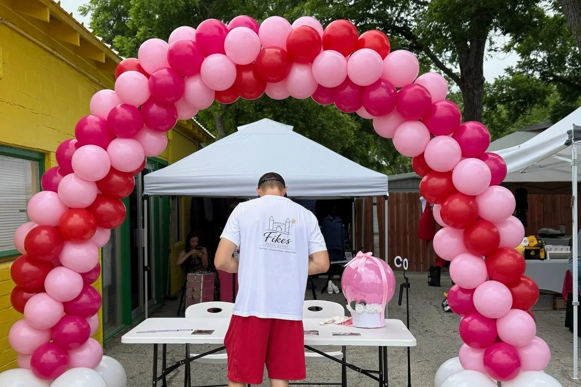 Pink and red balloon arch at an outdoor event, with a man in a white shirt and red shorts standing at a table underneath.