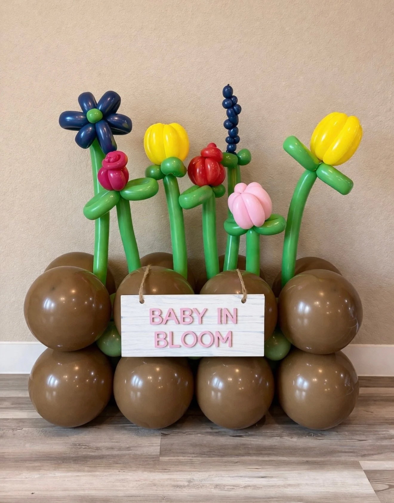 Balloon display featuring colorful balloon flowers on green balloon stems, arranged above a foundation of large brown balloons. A white sign with pink letters reads "Baby in Bloom."