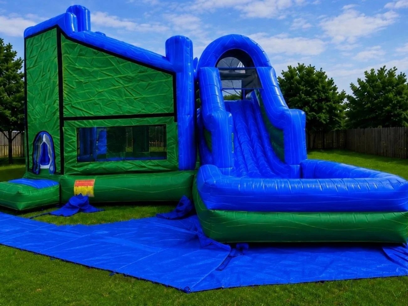 Blue and green inflatable water slide set up outdoors on grass with trees and a wooden fence in the background.