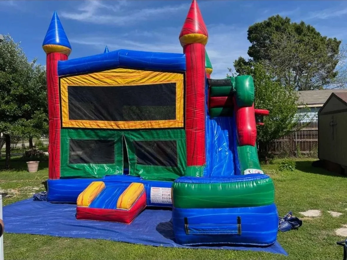 Colorful inflatable bounce house with a slide set up outdoors on green grass, surrounded by trees and a house in the background.