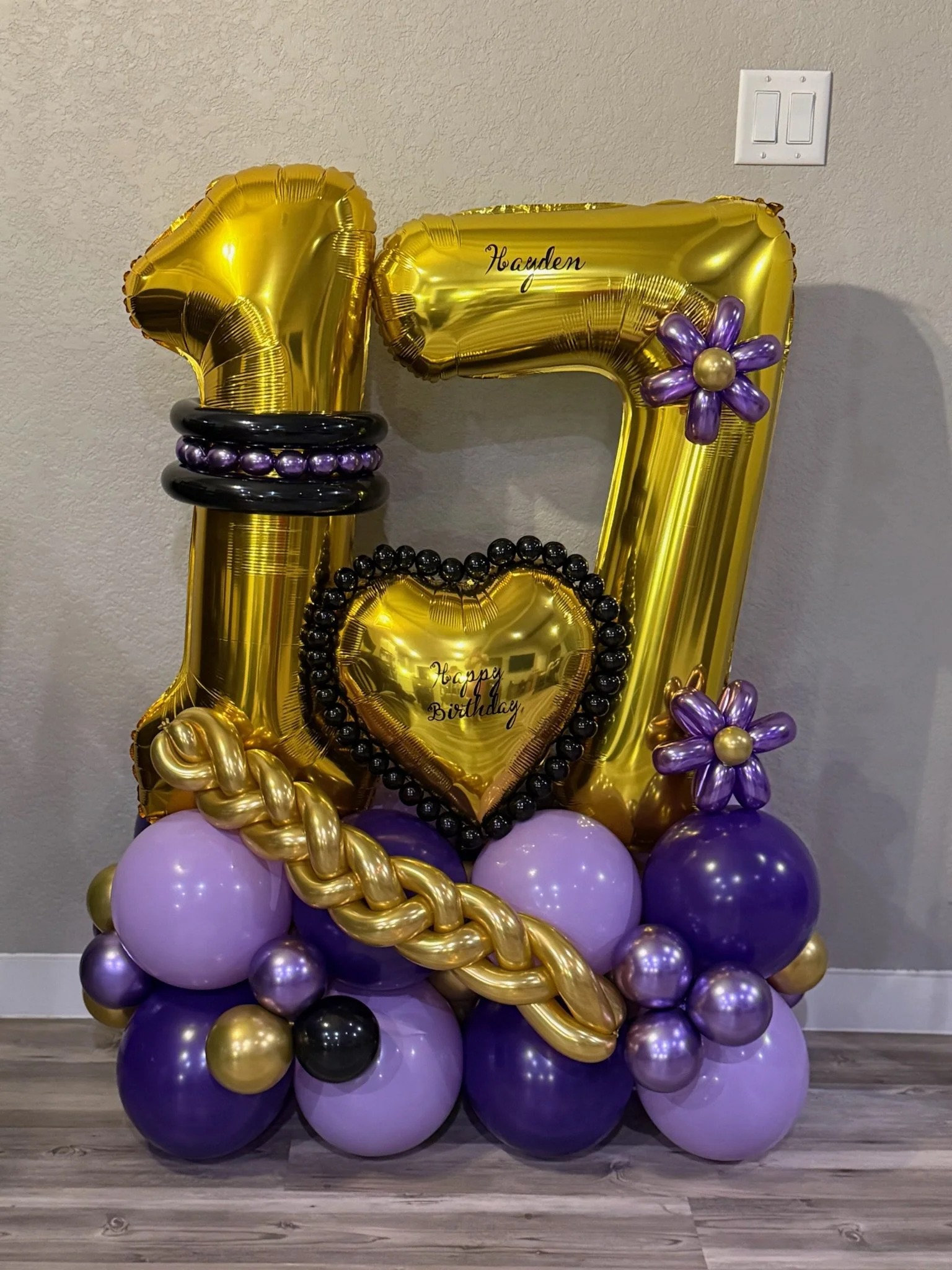 Yellow and purple birthday balloon decoration with large gold balloon numbers and purple flower accents, set on a cluster of purple, gold, and black balloons against a plain wall.