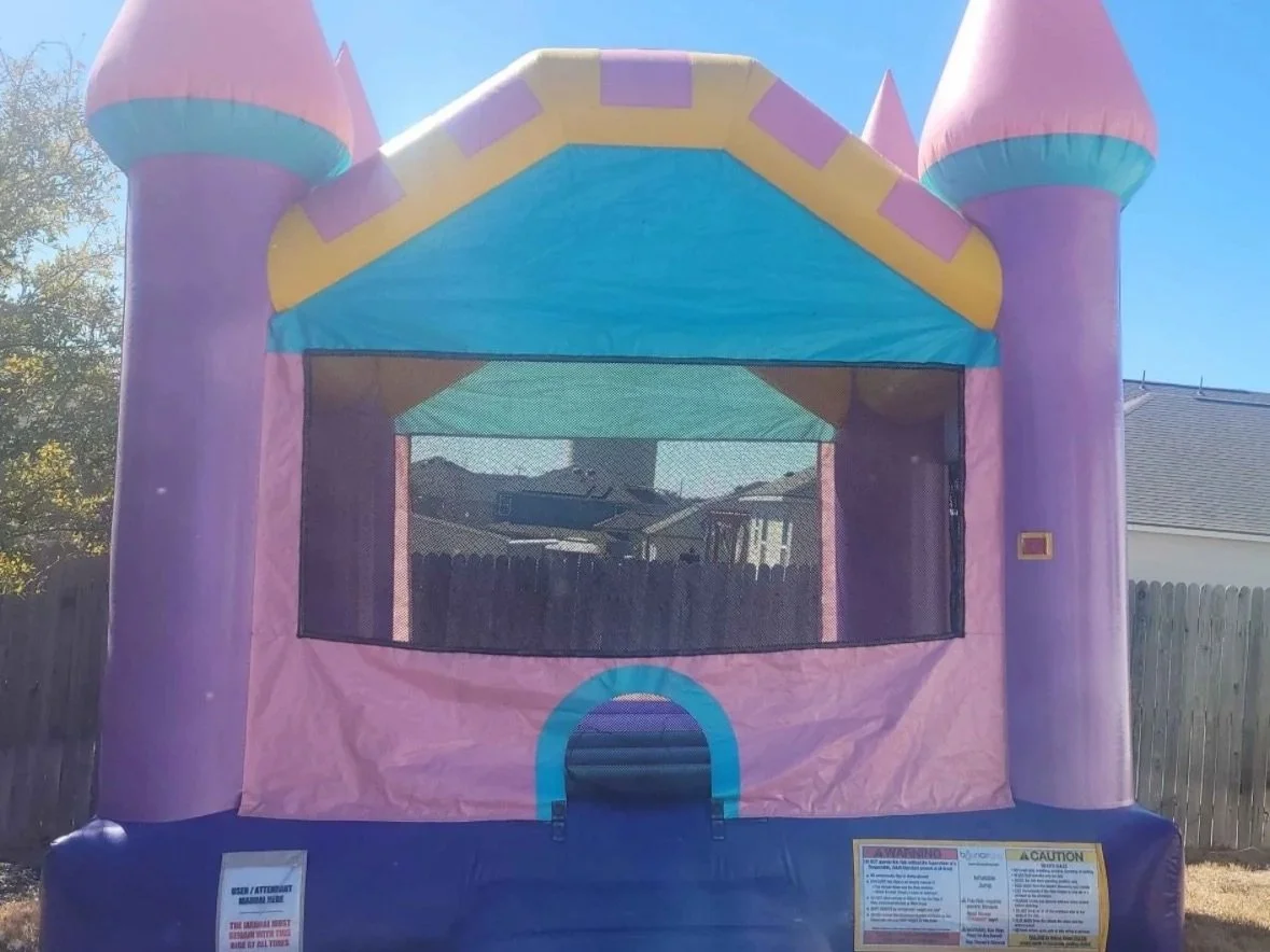 Colorful inflatable bounce house castle with purple, pink, yellow, and blue sections, set up outdoors on a grassy yard near a wooden fence on a sunny day.