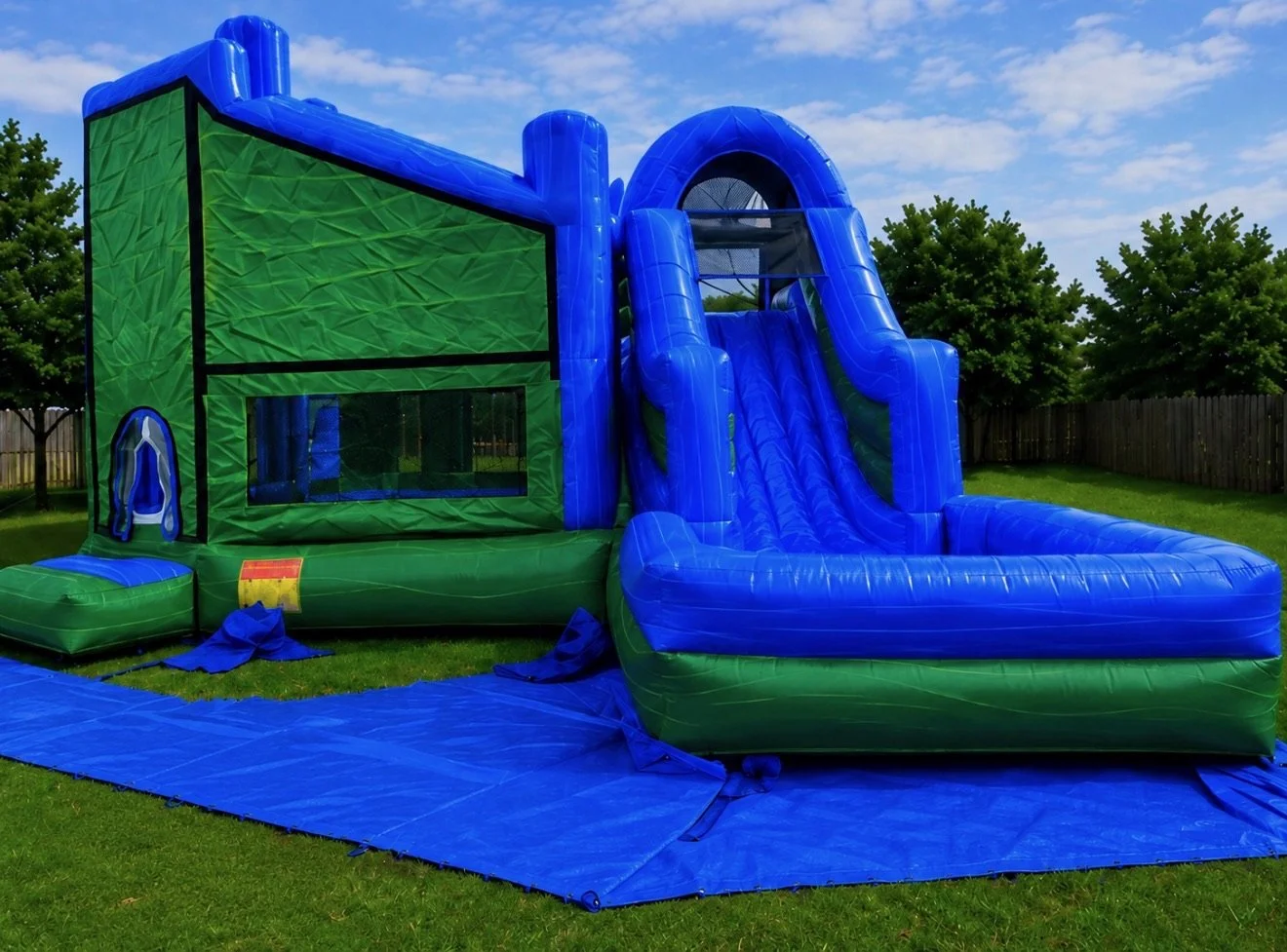 Colorful inflatable bounce house with water slide setup outdoors on grass, with trees and a wooden fence in the background.