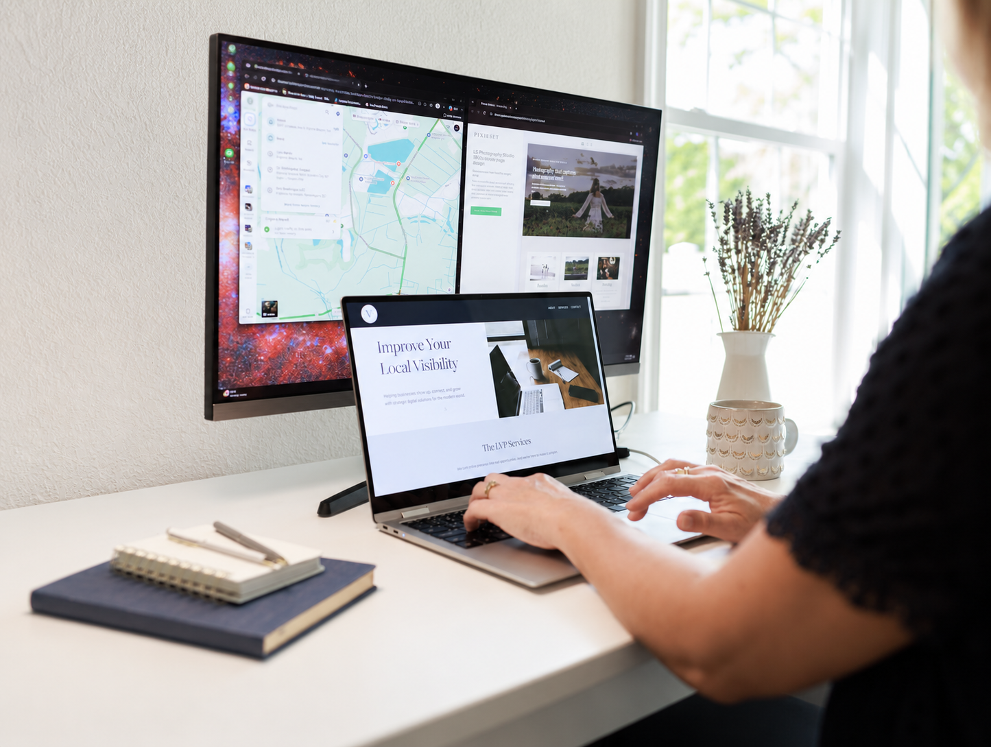 A person working at a white desk with a laptop and two monitor screens, with notebooks, a pen, a mug, and a vase of lavender in the background.