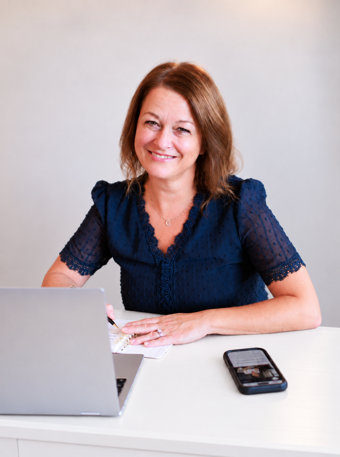 A woman with shoulder-length brown hair smiling, wearing a navy blue lace blouse, sitting at a white desk with a silver laptop, a smartphone, and a notebook.