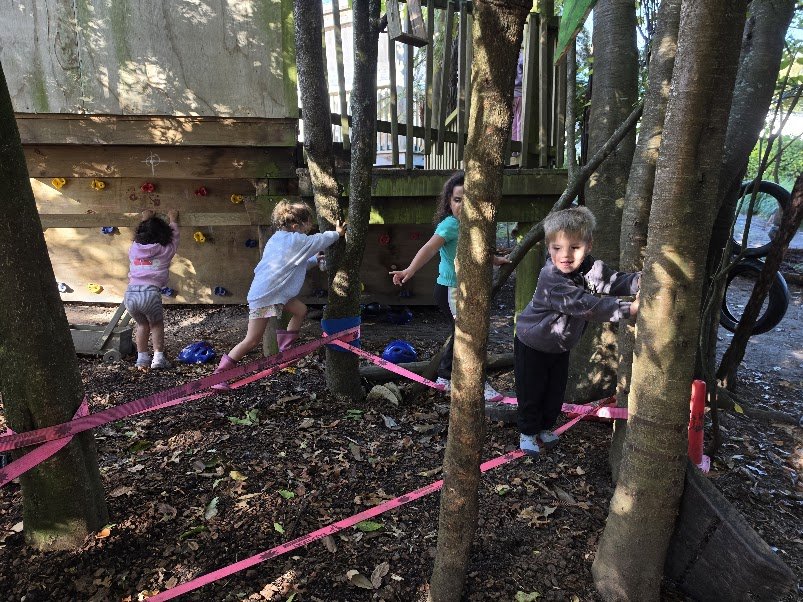 Four children playing outdoors among trees near a wooden climbing wall with colorful holds. Some children are climbing, others are waiting or watching.