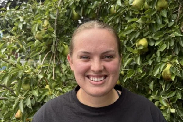 A smiling woman standing outdoors in front of a green bush with pears.