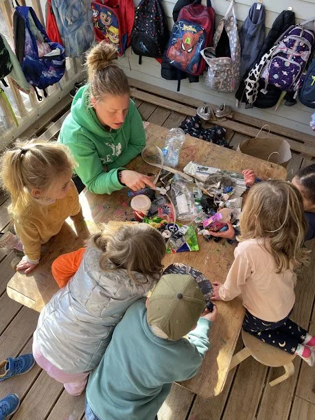 Group of children and an adult sitting around a table with various small objects and toys, in a room with backpacks hanging on the wall.
