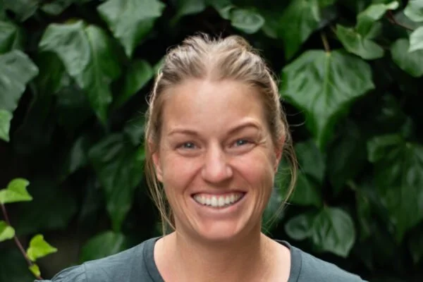 A woman smiling with tied-back blonde hair, standing in front of green leafy plants.