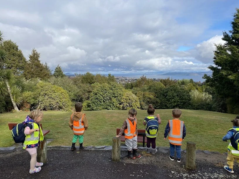 Group of children wearing bright safety vests standing outdoors on a grassy hilltop, overlooking a view of trees and a distant body of water.