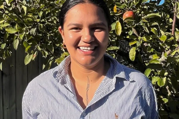 A smiling woman standing outdoors in front of an apple tree with apples on the branches.