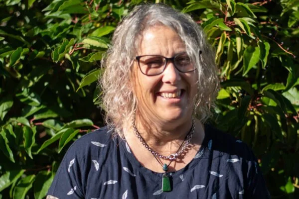 A smiling woman with curly gray hair and glasses outdoors surrounded by green foliage.