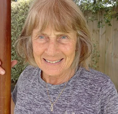 A smiling elderly woman with short, gray hair, wearing a gray shirt and a necklace, standing outdoors near trees and a wooden fence.