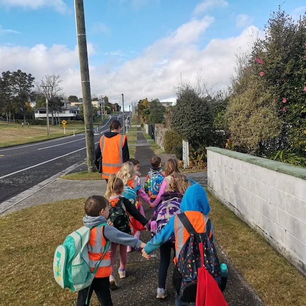 Children walking hand in hand on a sidewalk with an adult, all wearing colorful backpacks and safety vests, on a suburban street with houses, greenery, and cloudy sky.