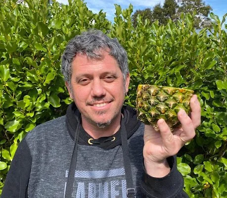 A man smiling outdoors holding a pineapple in front of green foliage.