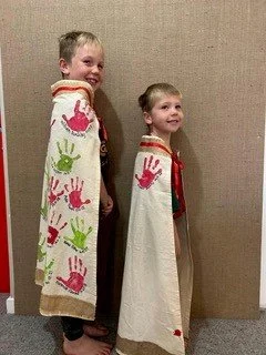 Two young boys standing against a beige wall, each wearing a white cape decorated with colorful handprints.