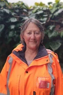 A woman outdoors wearing an orange safety jacket, standing in front of green plants.
