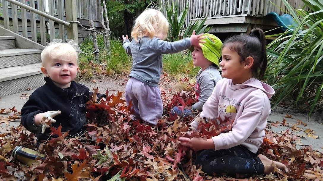 Three young girls and a young boy playing in a pile of autumn leaves outdoors near a wooden fence and steps.