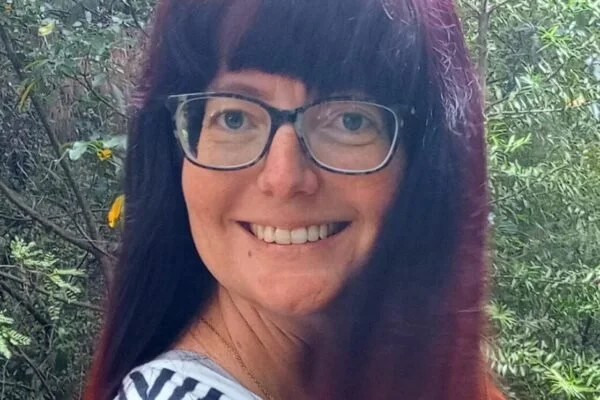 Close-up of a woman with long dark hair and glasses, smiling outdoors with green foliage in the background.