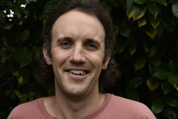 A man with curly brown hair and a wide smile standing outdoors in front of green leafy bushes.