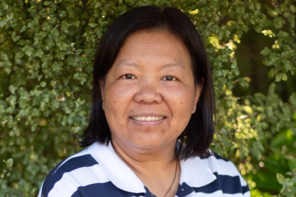 A woman smiling outdoors with green foliage in the background.
