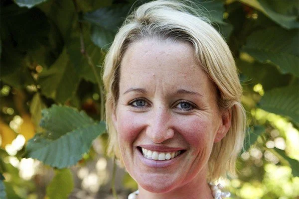 Close-up of a smiling woman with blonde hair outdoors surrounded by green leaves.
