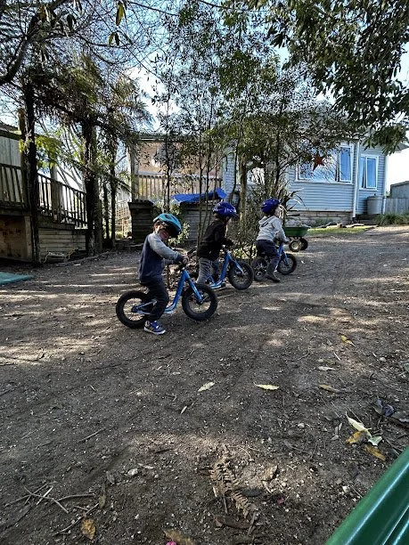 Three children wearing helmets riding balance bikes on a dirt path outdoors, with trees and a house in the background.