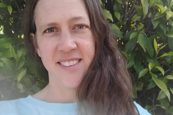 A woman with long brown hair and blue eyes smiling outdoors in front of lush green foliage.