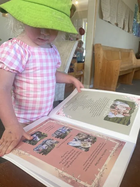 A young girl wearing a pink and white checkered shirt and a bright green sun hat, looking down at an open scrapbook or photo album on a table in a room with wooden furniture and hanging fabric or paper decorations.