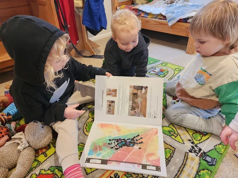 Three young children sitting on a colorful rug in a bedroom, looking at a book with pictures and colorful artwork.
